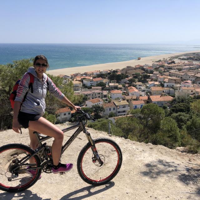 Photo d'une jeune fille sur son vélo avec en 2ème plan la plage et Leucate Plage vu d'en haut de la falaise